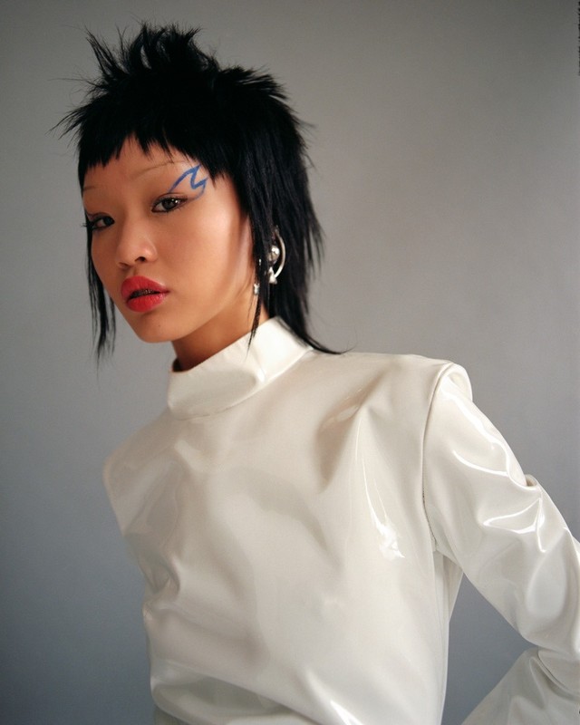 Woman in a white vinyl high-neck top poses against a grey background with bold blue makeup and a mullet hairstyle in a studio setting.
