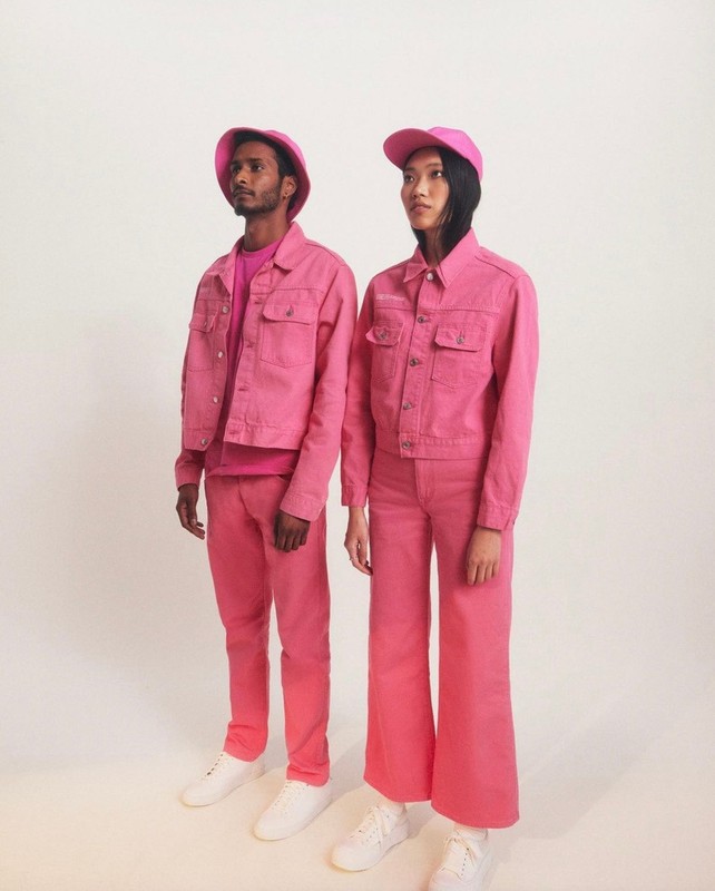 Two people in matching pink outfits, including jackets, pants, t-shirts, and hats, standing in a white studio with white sneakers.
