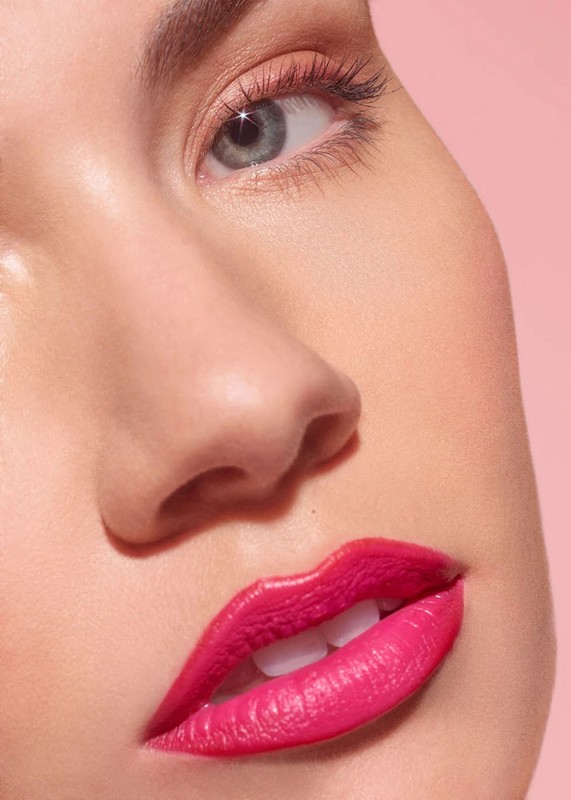 Close-up studio shot of a fair-skinned woman with bright pink lipstick, defined eyelashes, and light eyeshadow on a light pink background.
