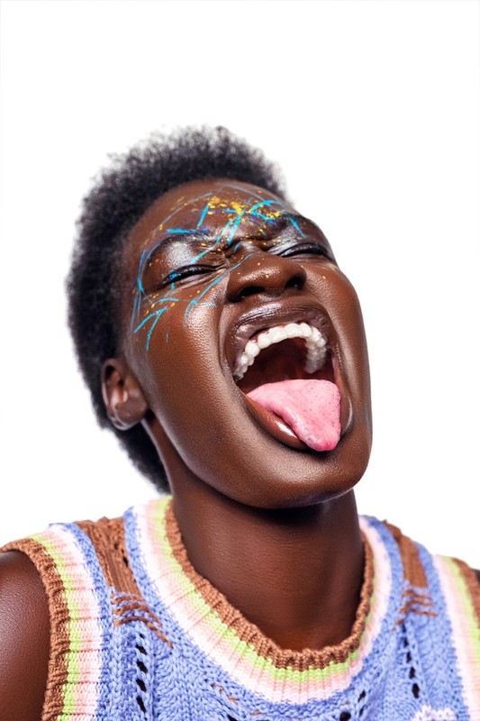 Close-up of a dark-skinned woman with face paint, eyes closed, and tongue sticking out on a white background. Fashion and beauty shot.