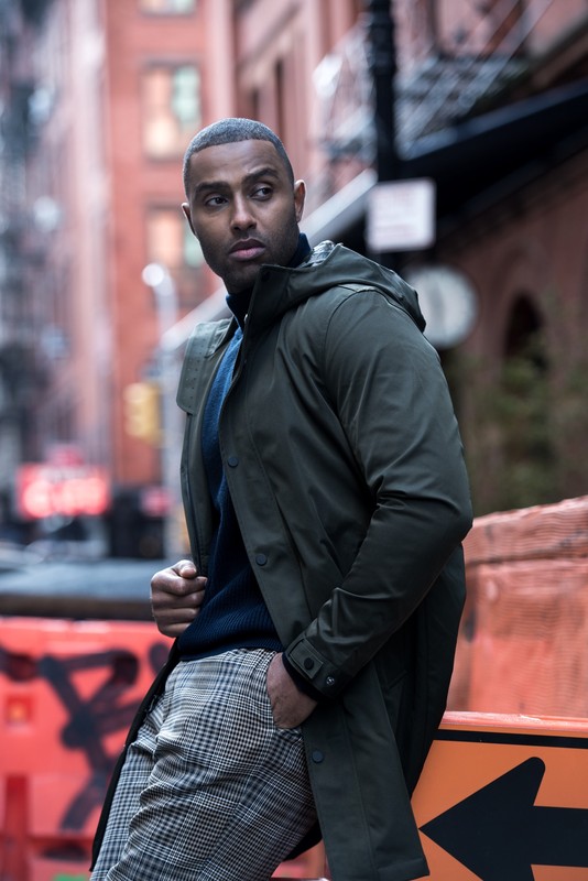 An adult male poses in an urban setting, wearing a dark green coat, a navy blue turtleneck sweater, and grey plaid pants, exuding urban fashion.