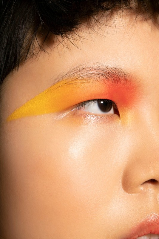 Close-up of a person's face featuring vibrant yellow and orange eyeshadow makeup, highlighting the eye and eyebrow.