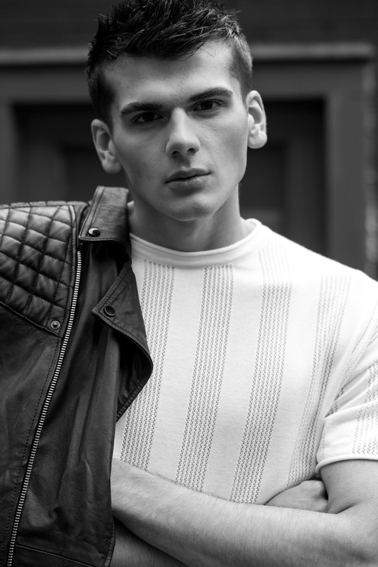 Monochrome portrait of a young adult male with a leather jacket draped over his shoulder and arms crossed looking at the camera.