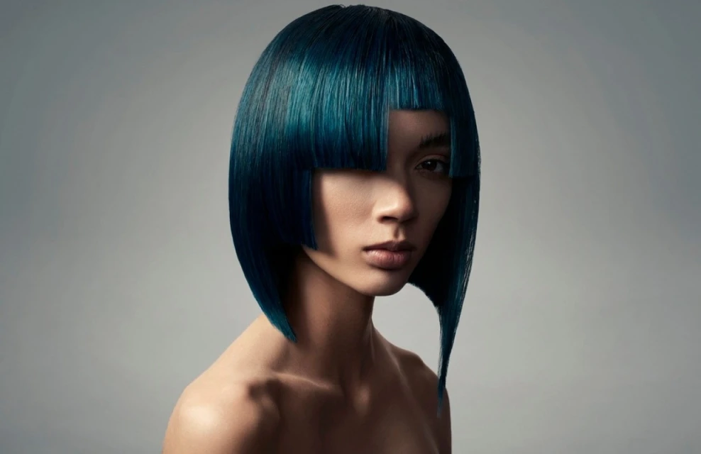 Close-up portrait of a young adult female with blue bob haircut and bare shoulders in front of a grey backdrop.