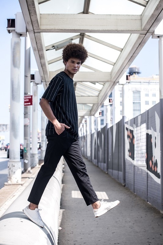 A young adult male model with afro, wearing a black and white striped shirt and black pants, poses on an elevated urban walkway.