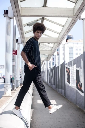A young adult male model with afro, wearing a black and white striped shirt and black pants, poses on an elevated urban walkway.