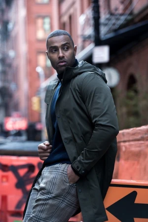 Man wearing a dark green hooded coat and plaid trousers posing outdoors with urban street background