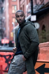 Man wearing a dark green hooded coat and plaid trousers posing outdoors with urban street background