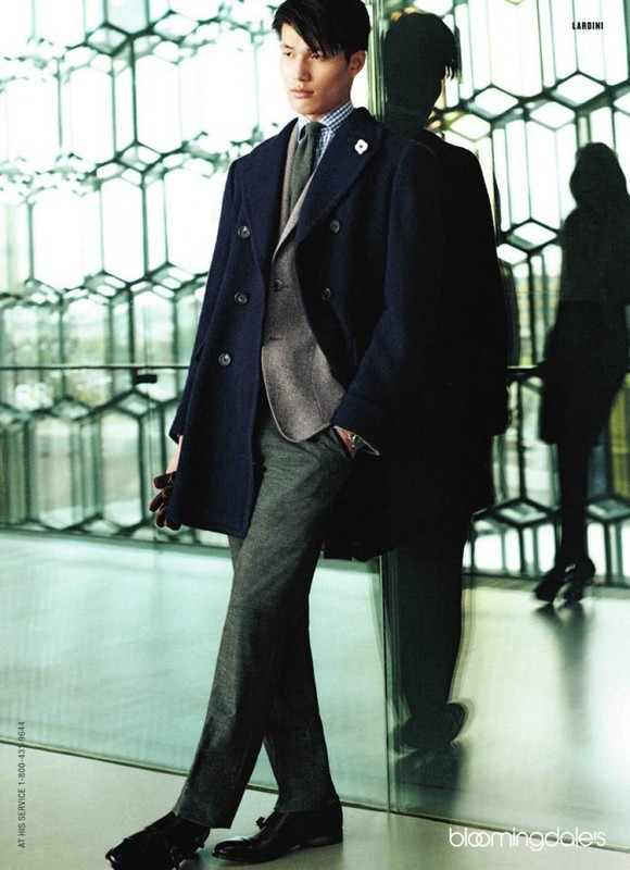 Man poses in a navy overcoat, grey suit, and grey trousers, standing next to a glass wall with a geometric honeycomb pattern in the background.