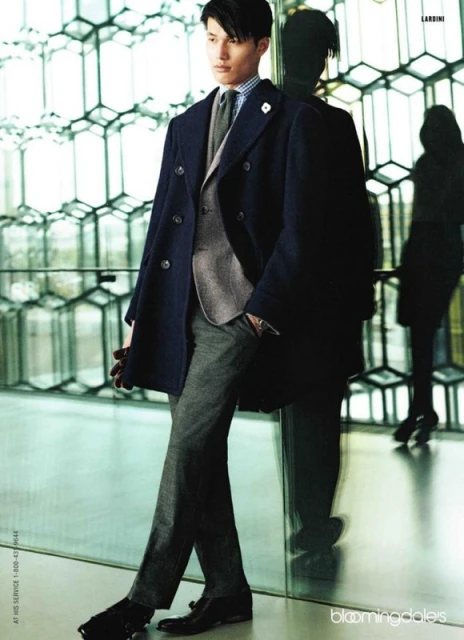 Man poses in a navy overcoat, grey suit, and grey trousers, standing next to a glass wall with a geometric honeycomb pattern in the background.