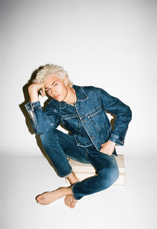 Young adult man with blonde hair poses in a denim jacket and jeans while sitting on a wooden block in a brightly lit photo studio.