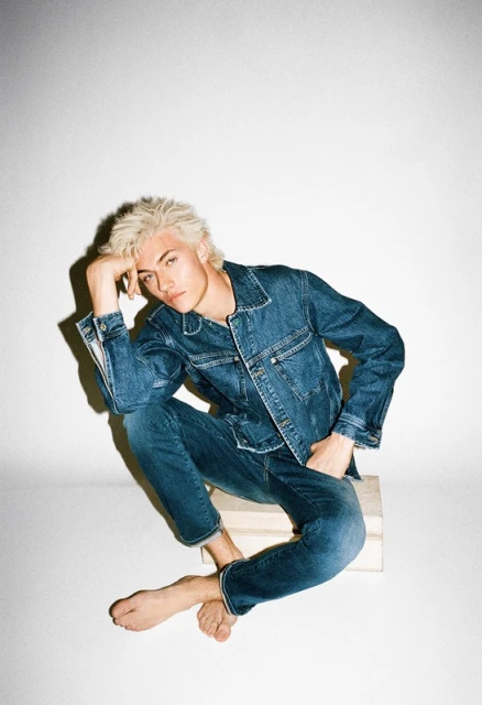 Young adult man with blonde hair poses in a denim jacket and jeans while sitting on a wooden block in a brightly lit photo studio.