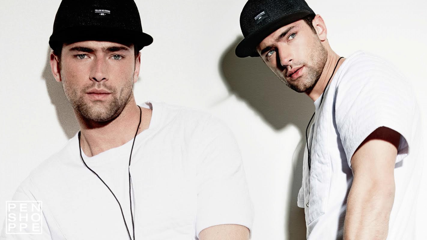 Two adult males wear black hats and white t-shirts in a studio shot against a white background. The men have similar features.