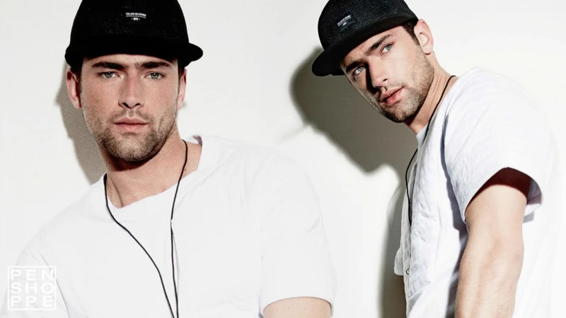 Two adult males wear black hats and white t-shirts in a studio shot against a white background. The men have similar features.