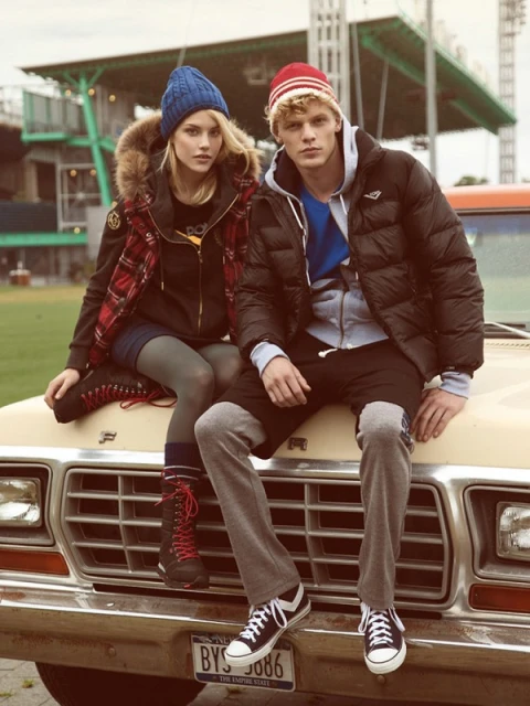 Two models, one male and one female, in winter clothing, sit on the hood of a vintage truck in front of a green outdoor space.