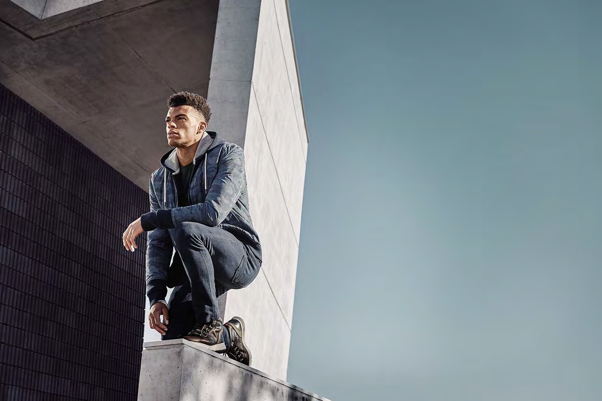 Athletic man crouching outdoors wearing a camo-patterned hoodie and dark jeans, next to a modern concrete architectural structure, sunny day.