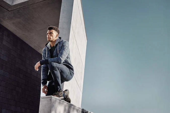Athletic man crouching outdoors wearing a camo-patterned hoodie and dark jeans, next to a modern concrete architectural structure, sunny day.