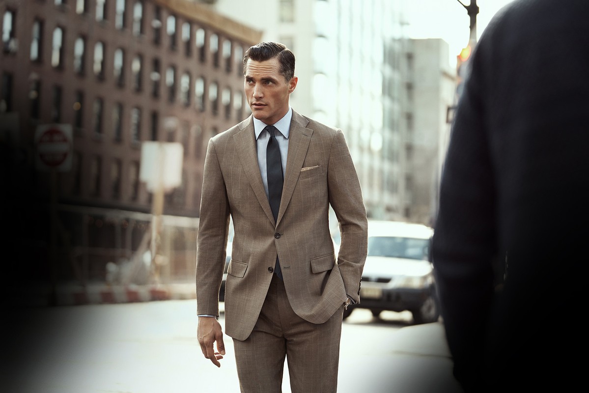 Man in tan suit walks on city street. Medium shot, outdoor setting, well-lit. Focus on subject; buildings and car in background.