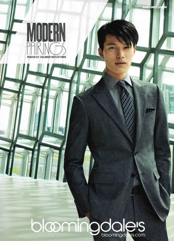 An Asian male fashion model poses indoors wearing a gray suit, tie, and dress shirt with a neutral facial expression near a window.