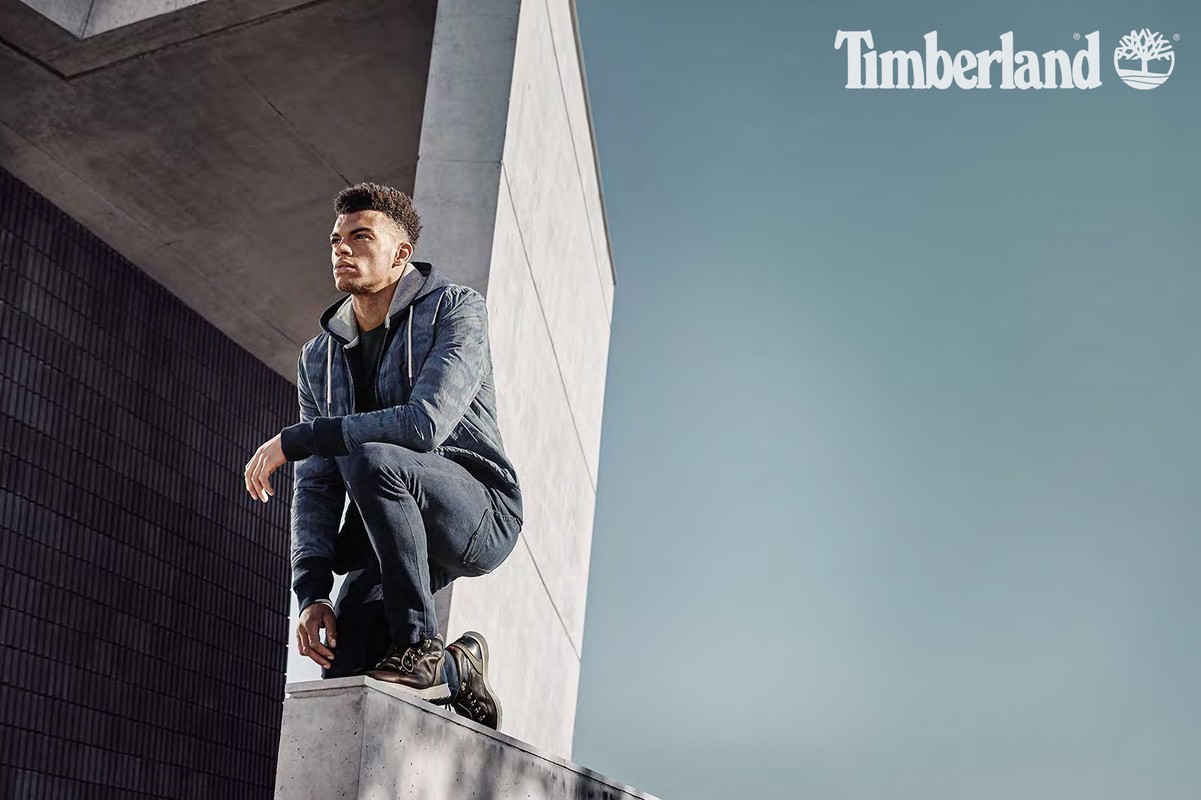 Adult male model squats on a concrete ledge in dark jeans and jacket for a Timberland advertisement outdoors, against a grey building.