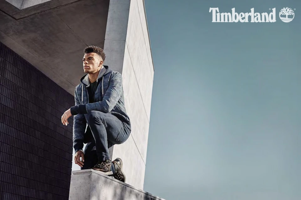 Adult male model squats on a concrete ledge in dark jeans and jacket for a Timberland advertisement outdoors, against a grey building.
