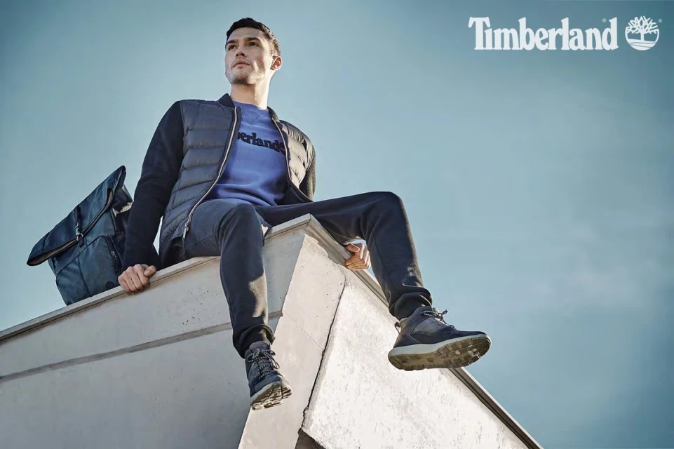 Man in Timberland apparel and boots sitting on a rooftop with a backpack against a clear blue sky, looking off-camera.