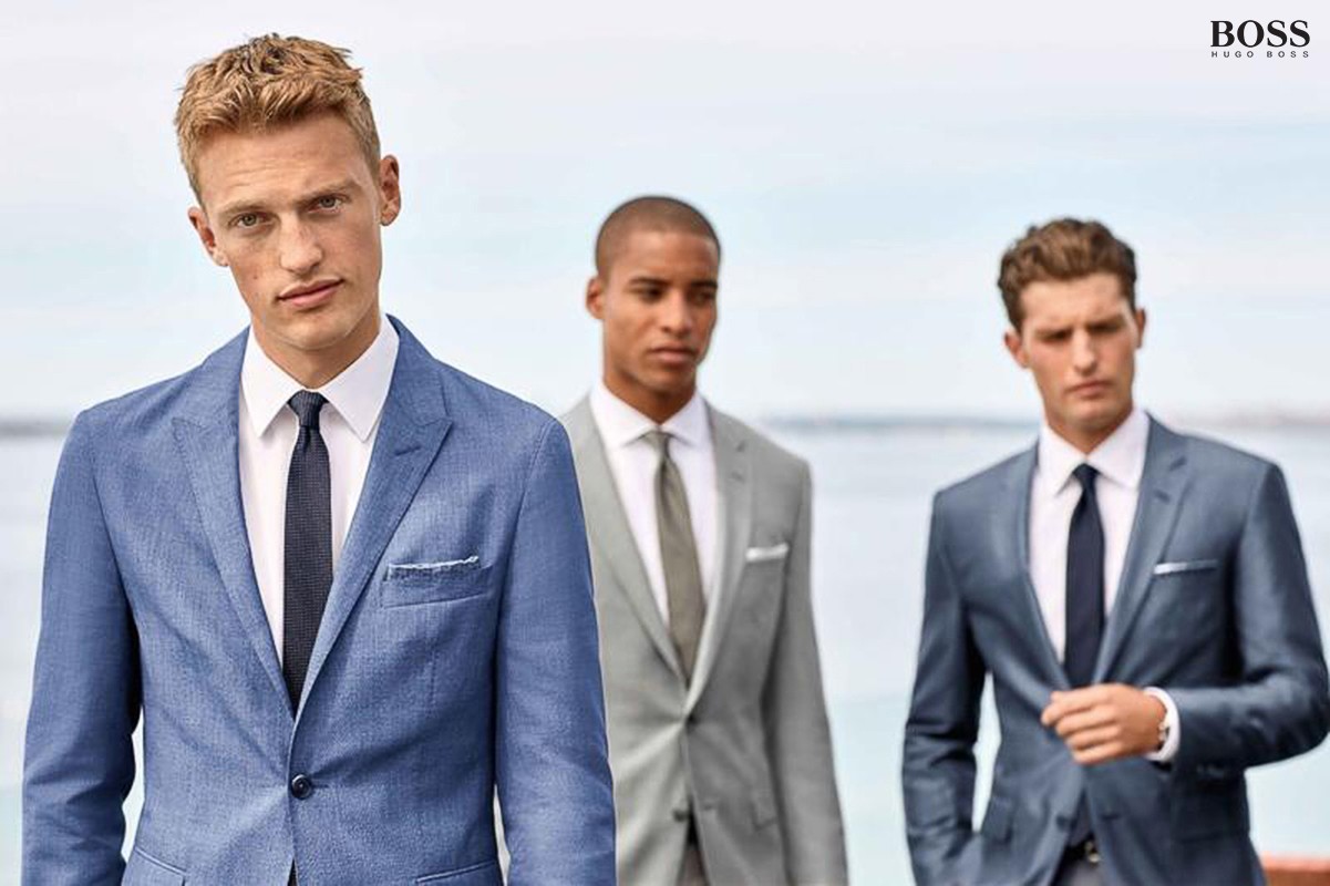 Three men are posing outdoors, wearing grey and blue suits with neckties. The BOSS logo is at the top, indicating a fashion advertisement.