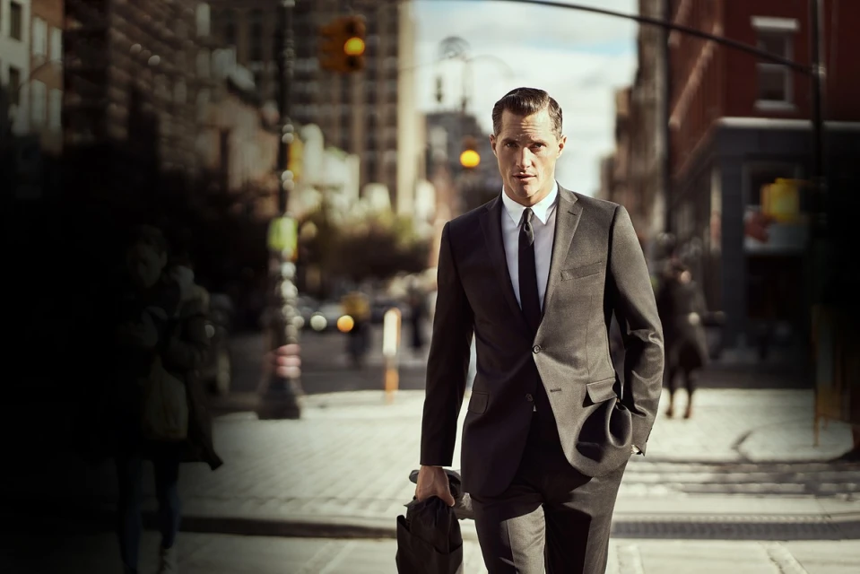 An adult male in a suit walks on a city street with buildings and a traffic light in the background, holding a briefcase.