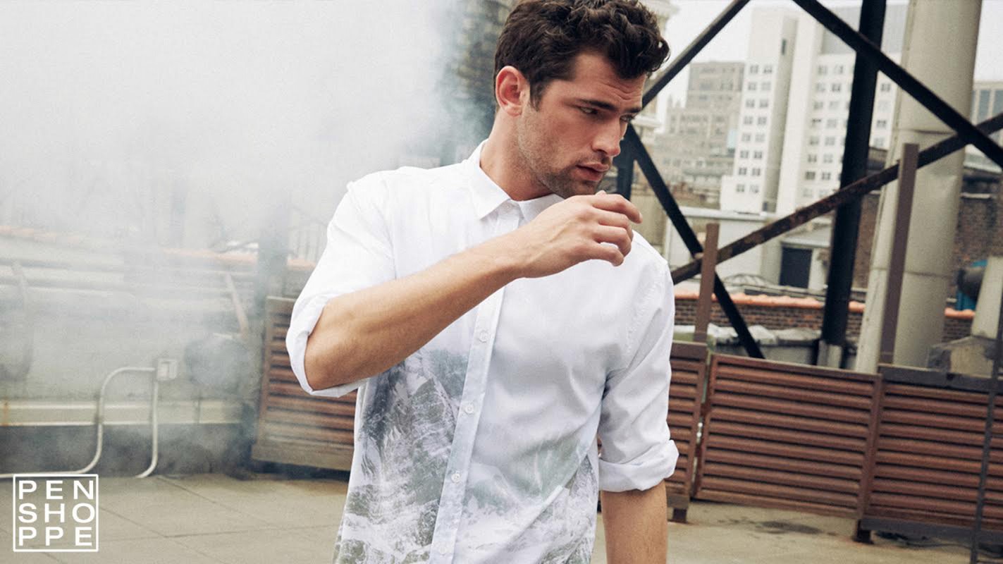 A casually posed adult male in a white button-down shirt stands on a rooftop with an urban background and diffused, natural lighting.