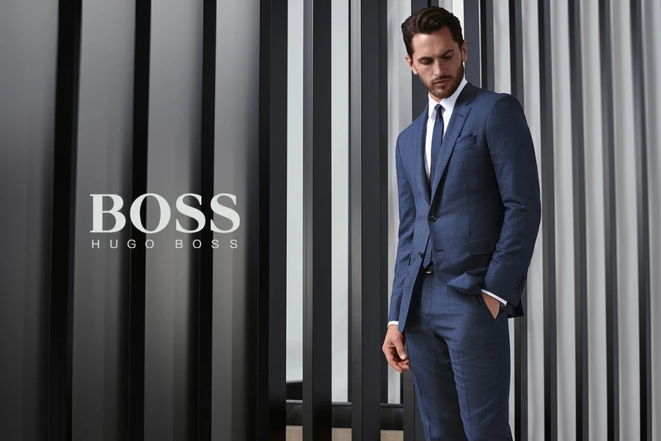 A stylish adult male poses in a blue suit in front of a vertically striped grey and black background, showcasing fashion apparel.