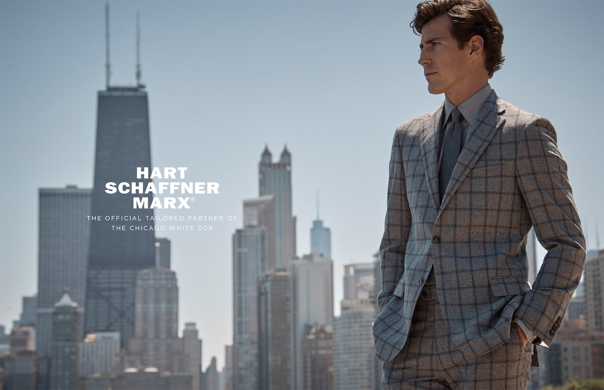 Adult male fashion model poses in a plaid suit with a cityscape backdrop on a sunny day for an outdoor advertising campaign.