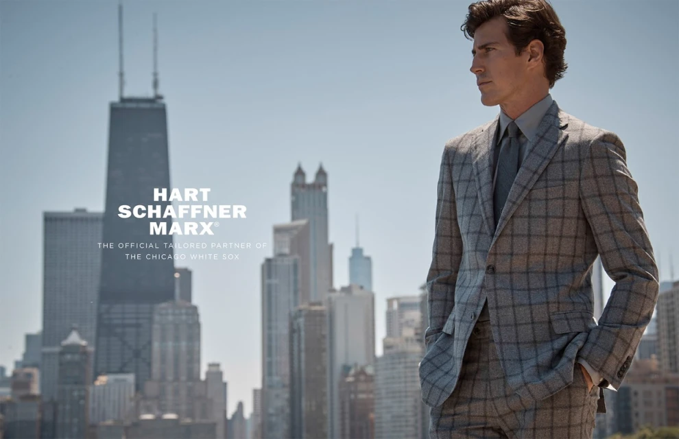 Adult male fashion model poses in a plaid suit with a cityscape backdrop on a sunny day for an outdoor advertising campaign.