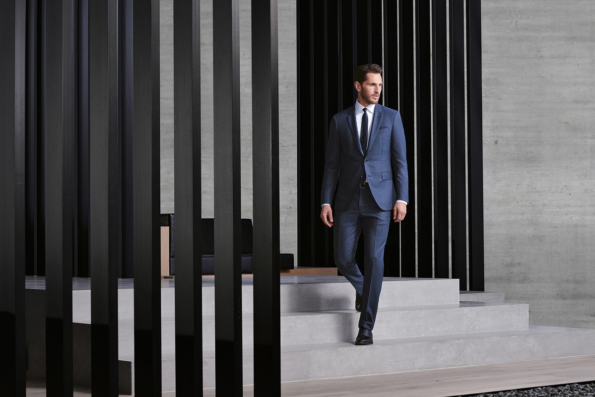 Man descends stairs in a blue suit, white shirt, and dark tie. Architectural interior featuring concrete and dark wood slats.