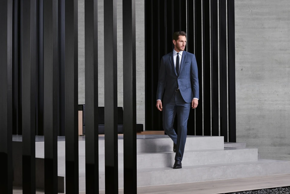 Man descends stairs in a blue suit, white shirt, and dark tie. Architectural interior featuring concrete and dark wood slats.