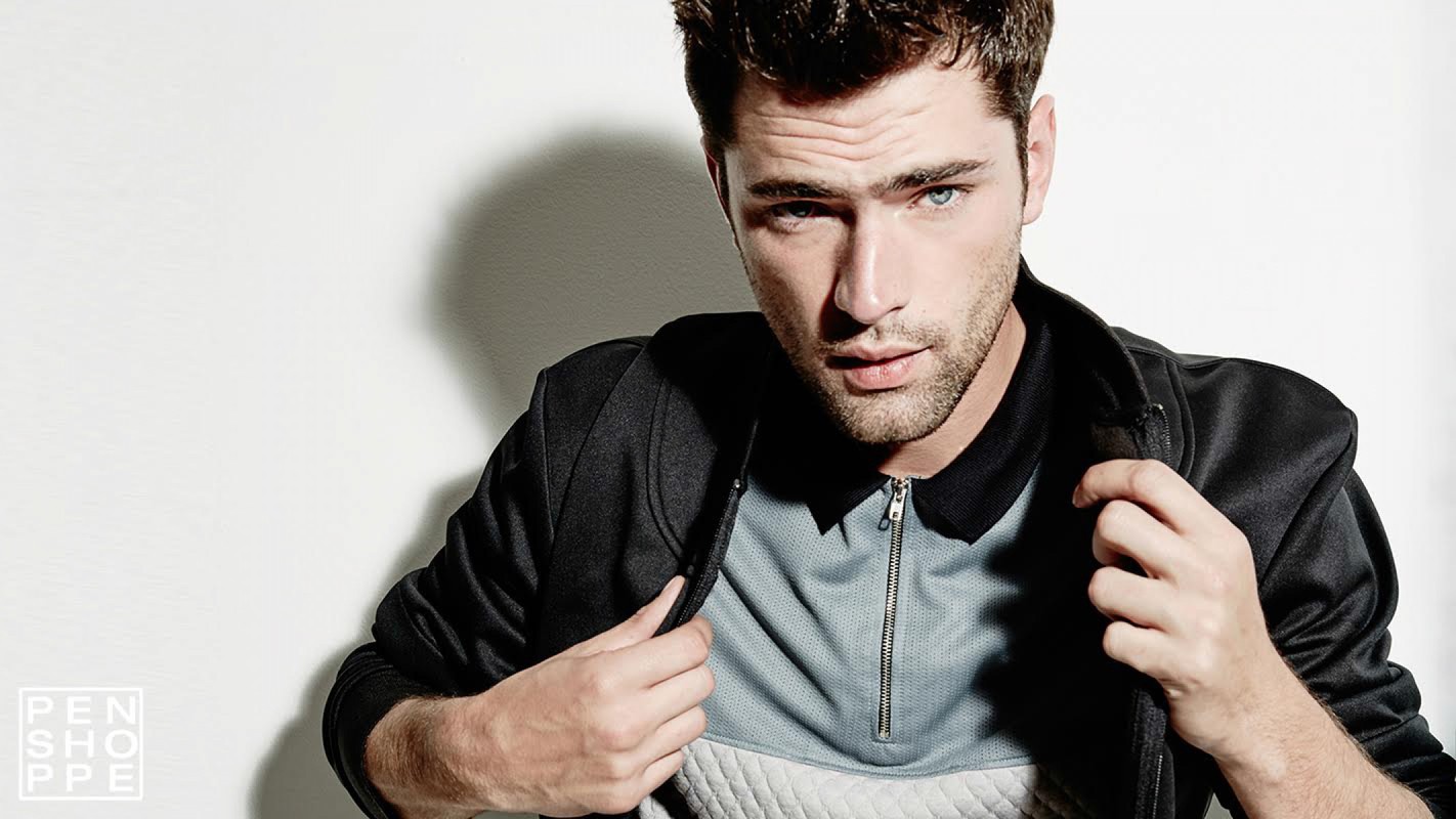 Close-up portrait of a handsome adult male model with blue eyes, wearing a black jacket and grey shirt, against a white backdrop.