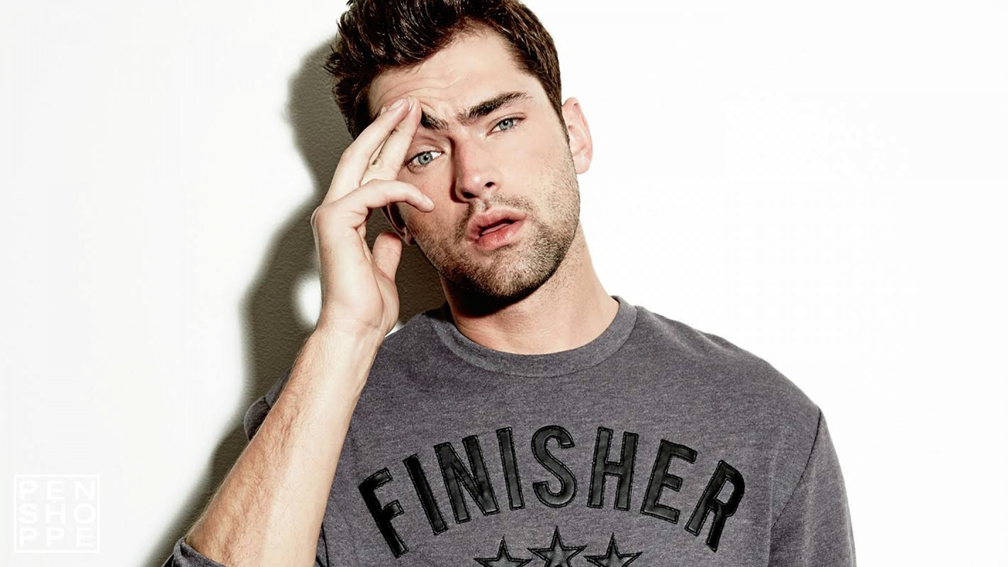 Close-up studio shot of a young adult male model with hand near his face, wearing a gray long-sleeved shirt against a white background.