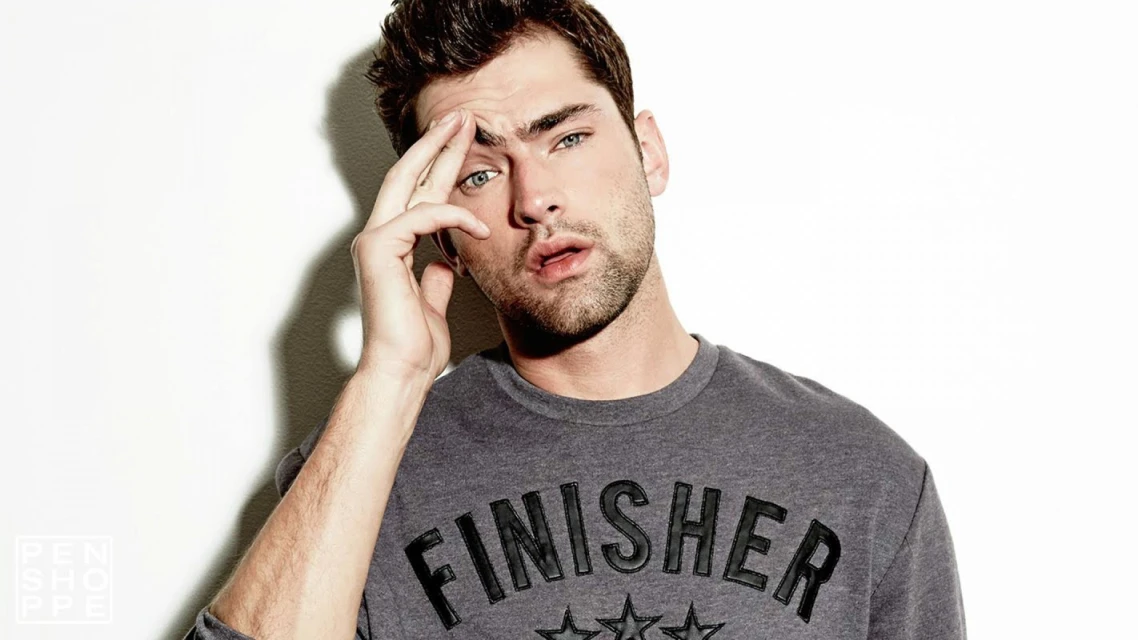 Close-up studio shot of a young adult male model with hand near his face, wearing a gray long-sleeved shirt against a white background.