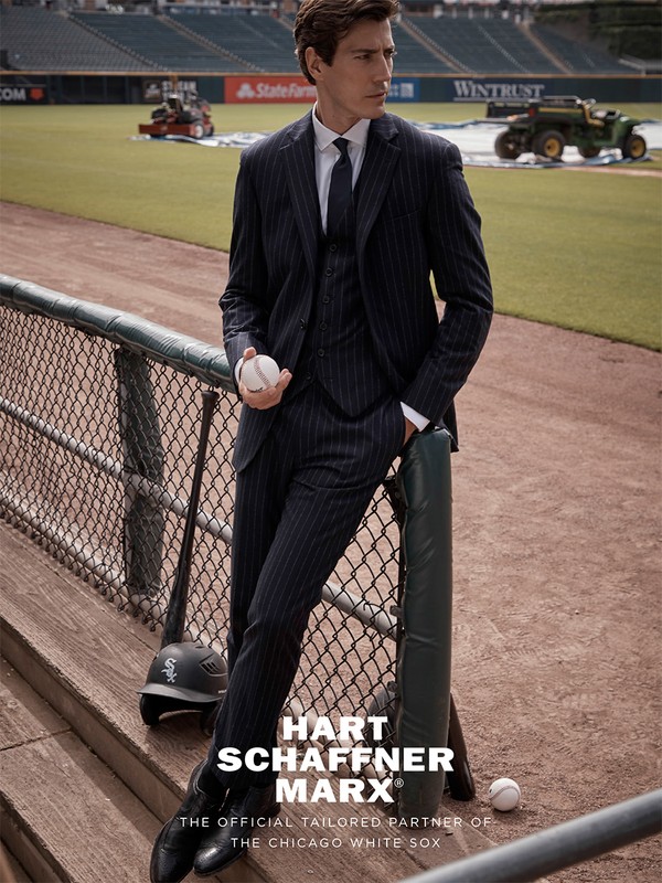 Man in pinstripe suit poses on baseball field near fence, holding ball with helmet and field visible in the background in daytime.