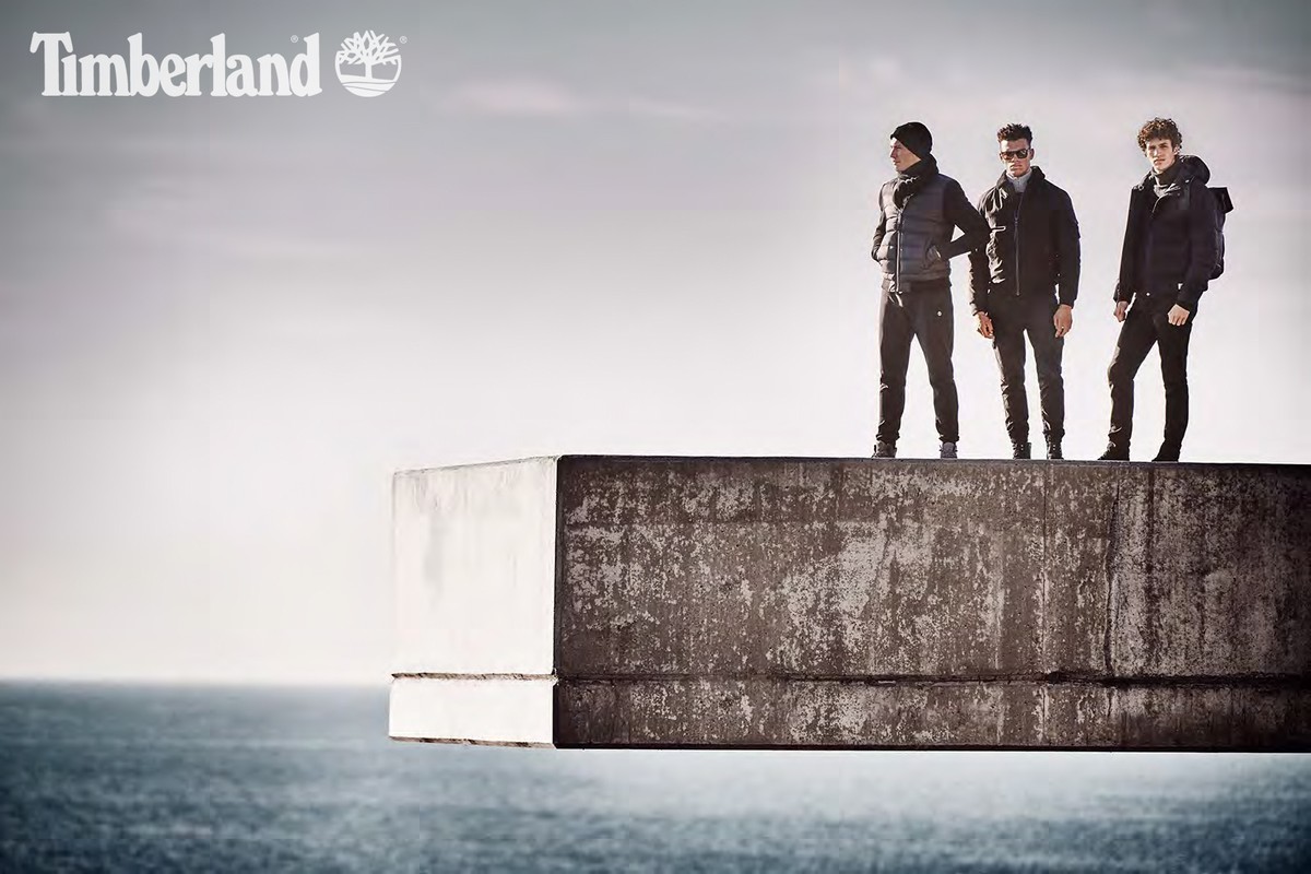 Three men in outdoor apparel stand on a concrete structure overlooking the ocean under an overcast sky, promoting the Timberland brand.