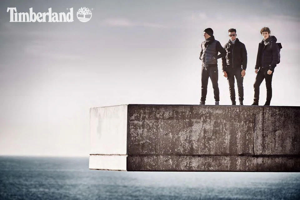 Three men in outdoor apparel stand on a concrete structure overlooking the ocean under an overcast sky, promoting the Timberland brand.