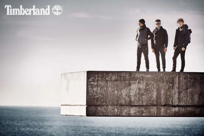 Three men in outdoor apparel stand on a concrete structure overlooking the ocean under an overcast sky, promoting the Timberland brand.