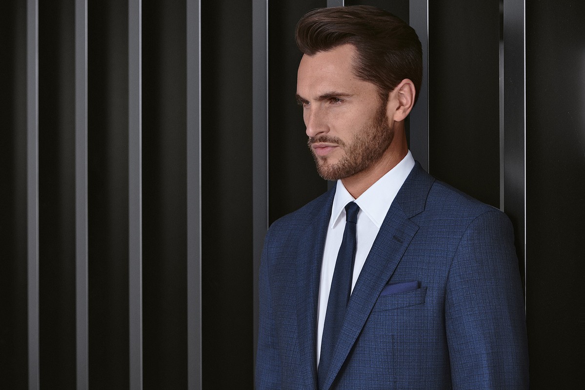Headshot of an adult male wearing a blue suit, white dress shirt, and blue tie posing in front of a black slatted background.