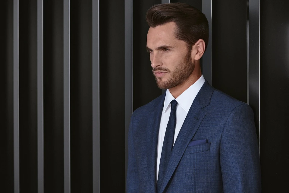 Headshot of an adult male wearing a blue suit, white dress shirt, and blue tie posing in front of a black slatted background.