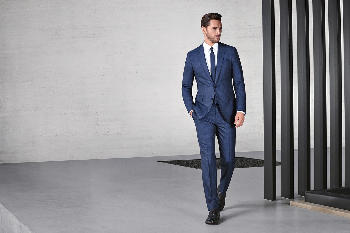 A stylish adult male model poses in a blue suit, white dress shirt, and blue tie in an indoor setting with modern architecture.