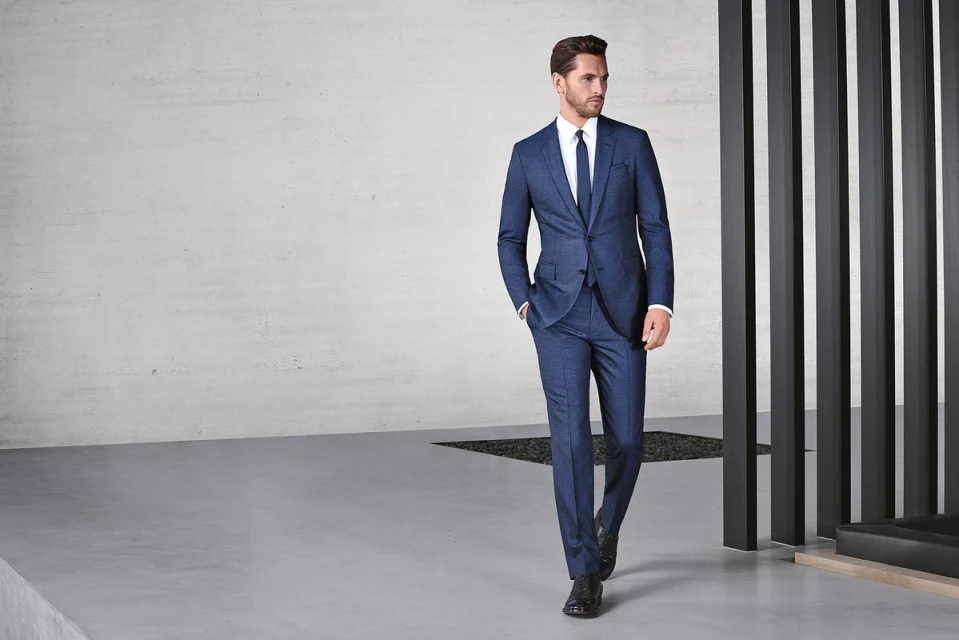 A stylish adult male model poses in a blue suit, white dress shirt, and blue tie in an indoor setting with modern architecture.