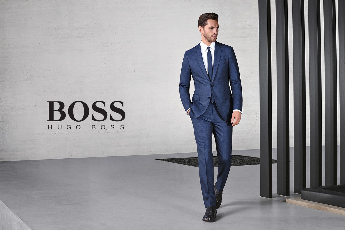 Adult male in a blue suit and tie walks in front of a modern wall with BOSS branding, showcasing fashion and style in a studio.