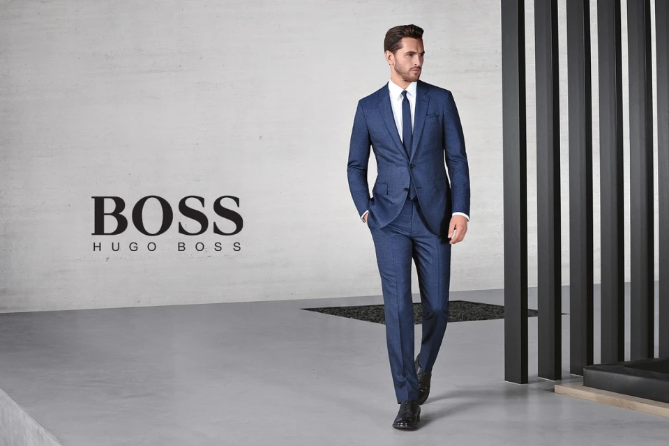 Adult male in a blue suit and tie walks in front of a modern wall with BOSS branding, showcasing fashion and style in a studio.