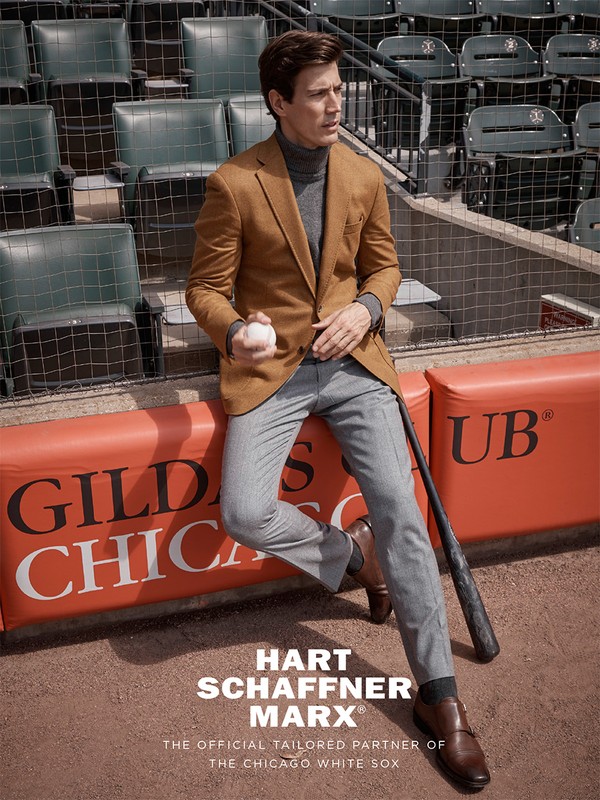 Male model in a sports stadium wearing a tan blazer and gray pants, holding a baseball with a baseball bat on the ground.