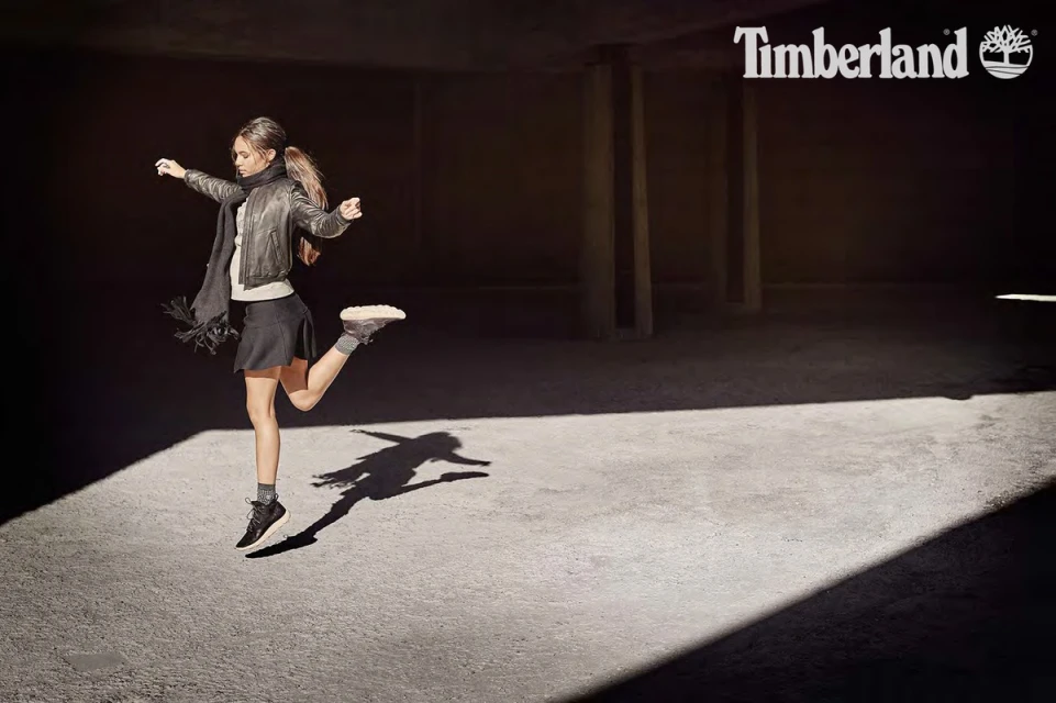 A young woman jumps mid-air in a stylish outfit: black skirt, jacket, and athletic shoes, against a shadowy urban concrete background.