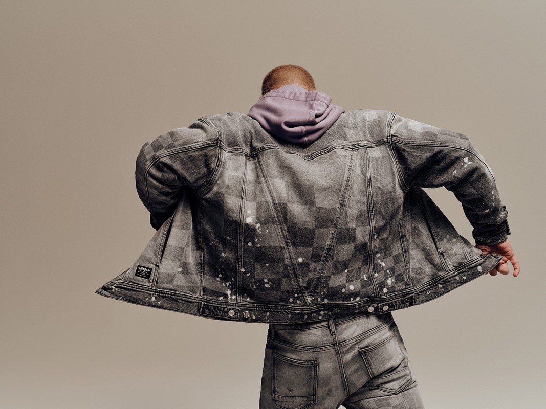 Person wearing a gray, checkerboard denim jacket with a lilac hoodie, standing against a neutral background in a studio setting.